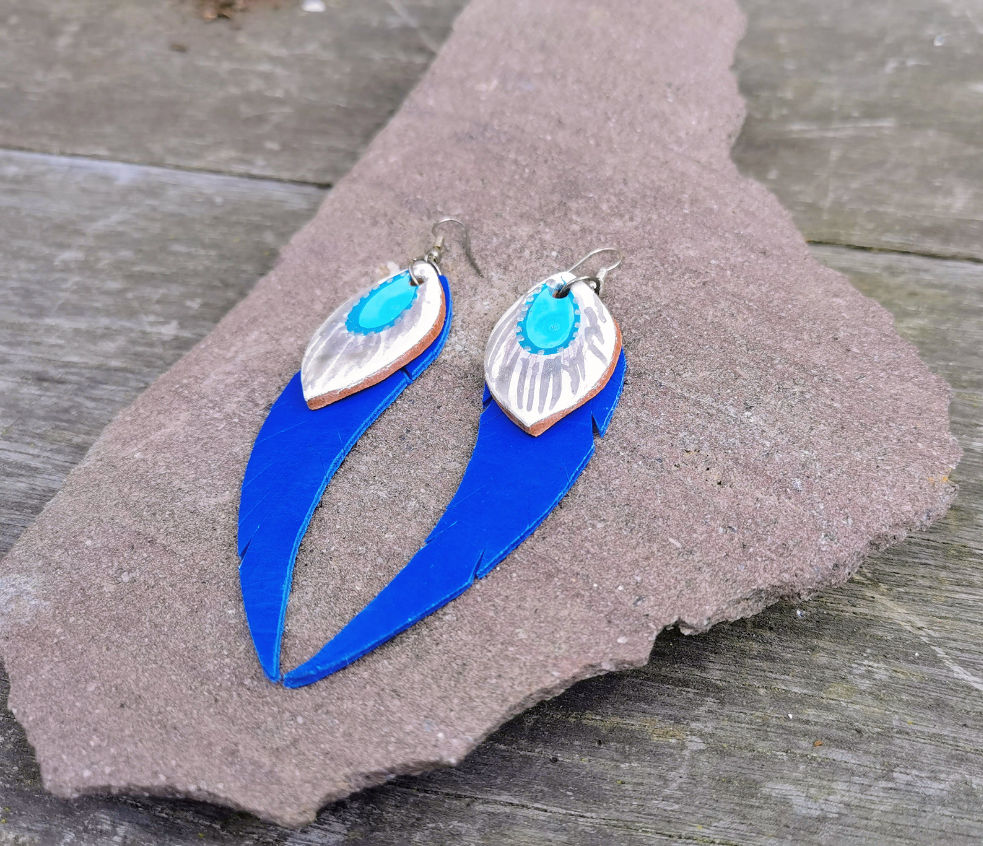 A pair of blue and white hand painted leather dangle earrings laid on a textured surface.