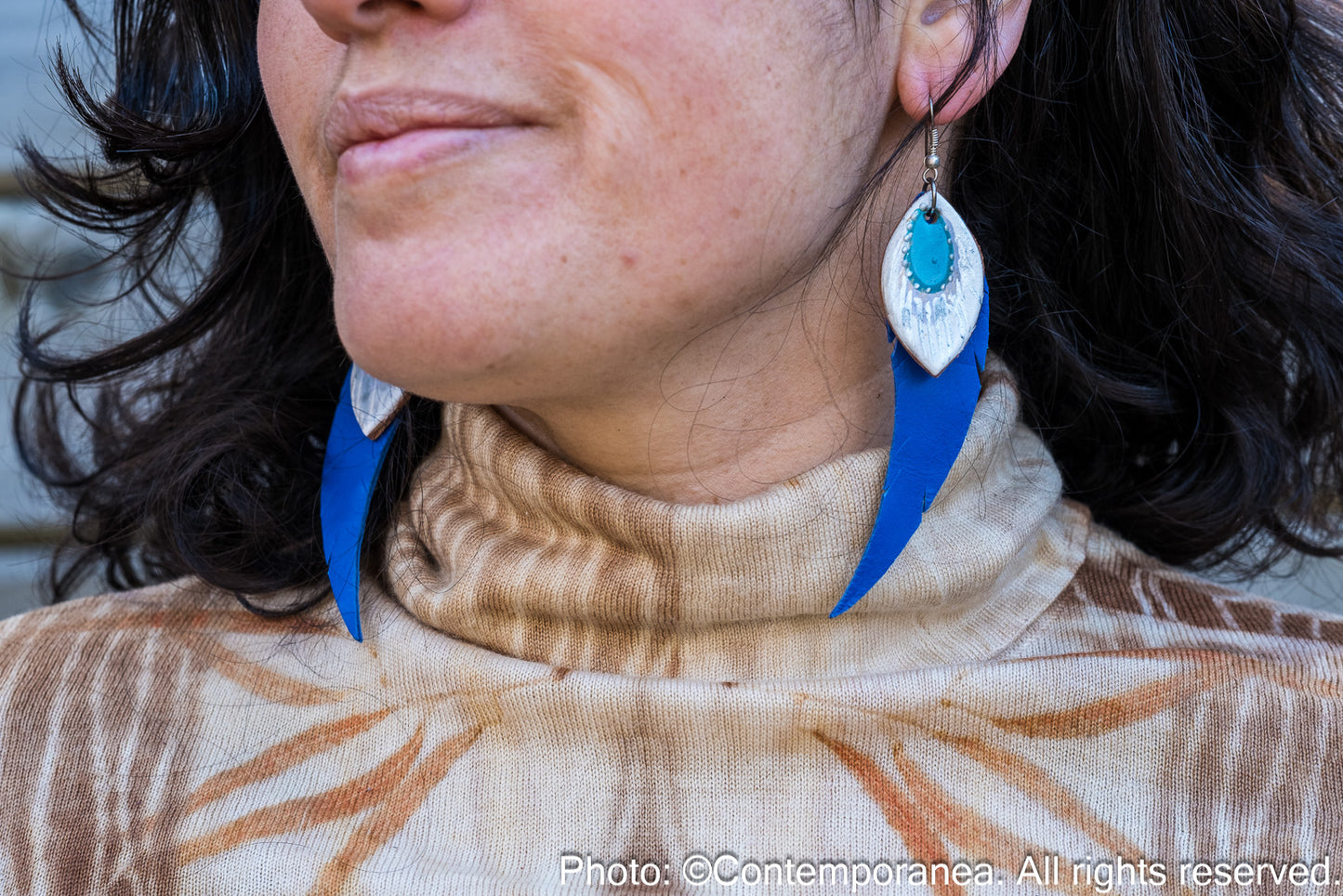 Close-up of a woman wearing a patterned top and feather-shaped blue and silver earrings against a wooden background