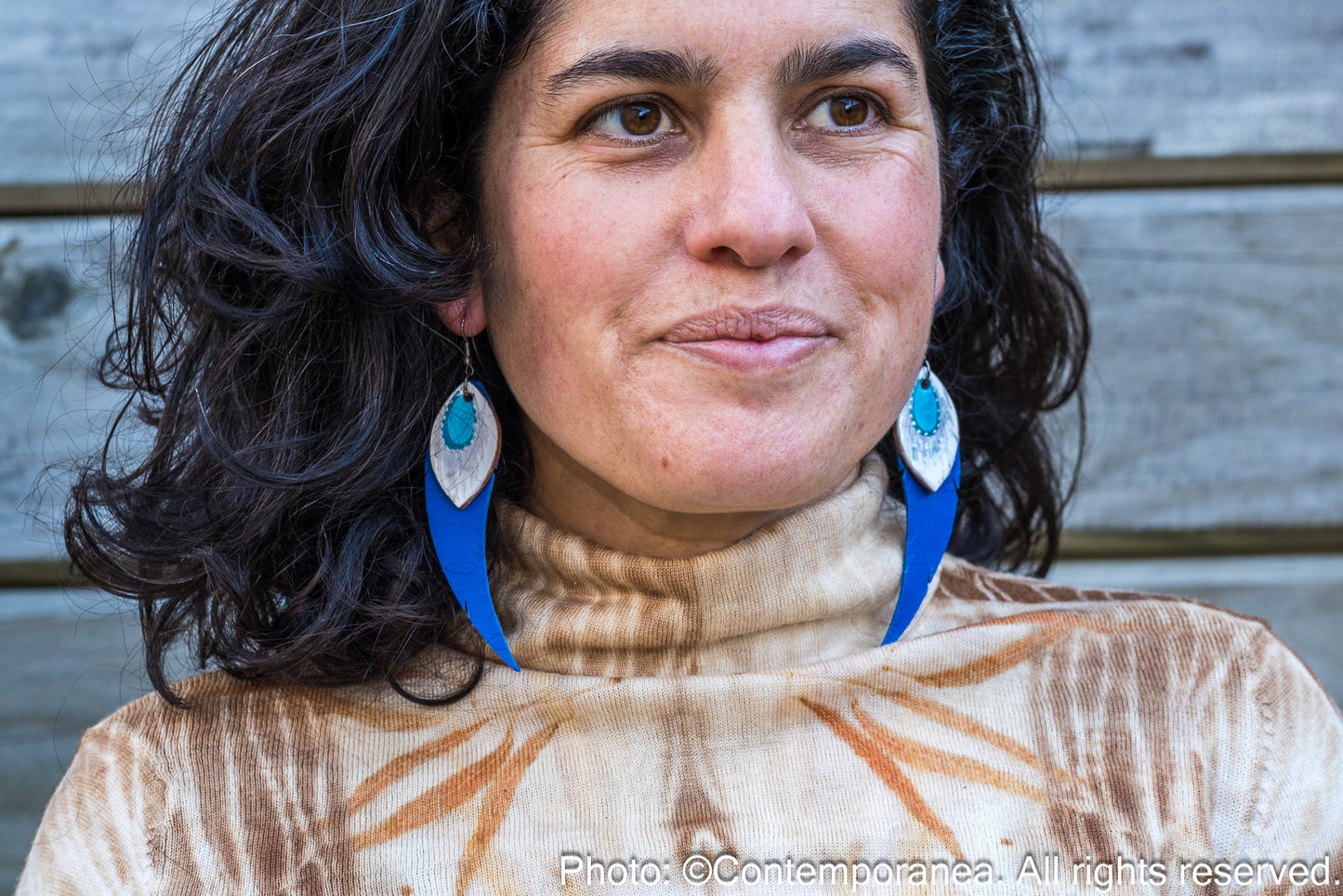 Woman wearing a patterned top and feather-shaped blue and silver earrings against a wooden background
