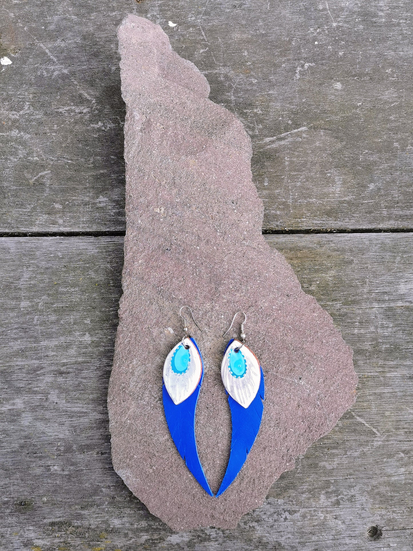 A pair of blue and white hand painted leather dangle earrings laid on a textured surface.