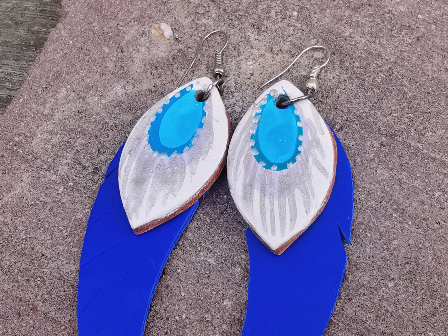 Close-up of a pair of blue and white hand painted leather dangle earrings laid on a textured surface.