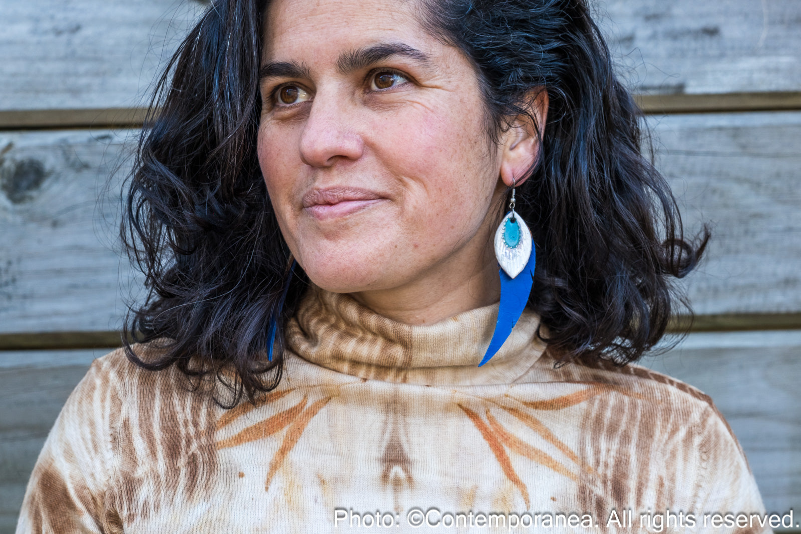 Woman wearing a patterned top and feather-shaped blue and silver earrings against a wooden background